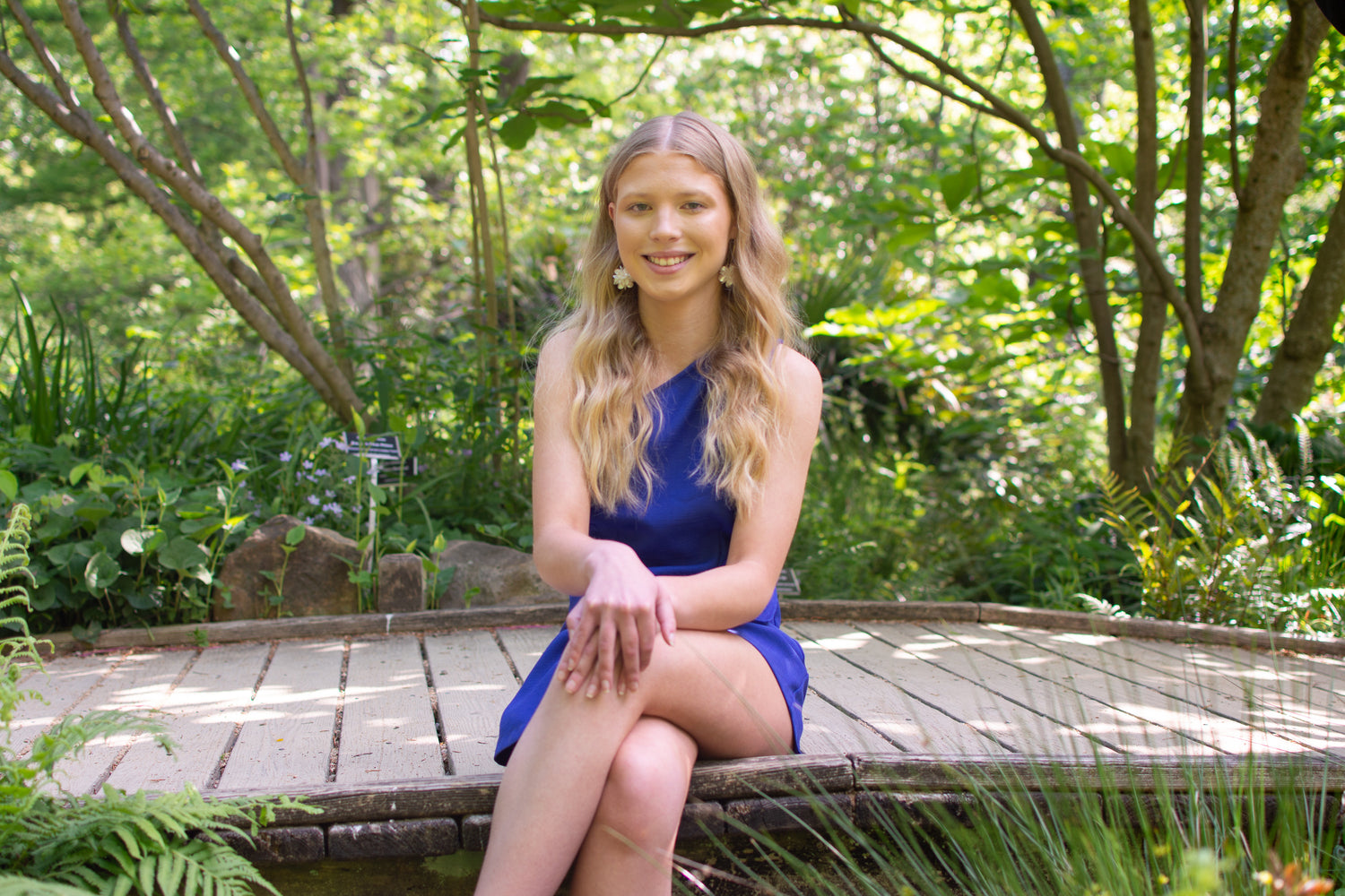 Rockstar Beaded Jewels' owner and artist, Lacey-Rae, wearing a blue dress sitting on a bridge surrounded by greenery